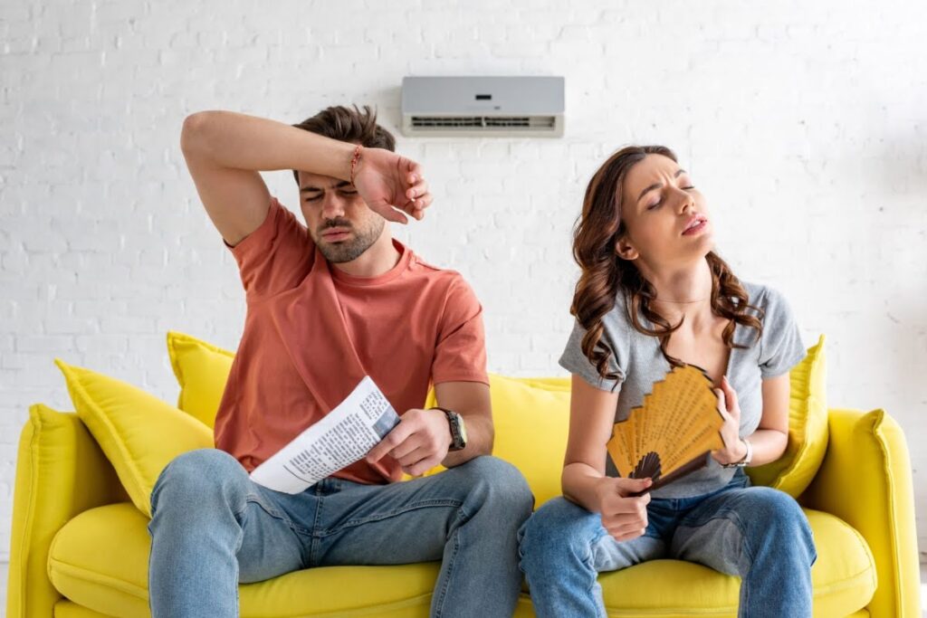 A husband and wife sitting on a bright yellow couch under an AC unit that is blowing hot air. The man is wiping his arm across his sweaty forehead and the woman is fanning herself with a paper fan.